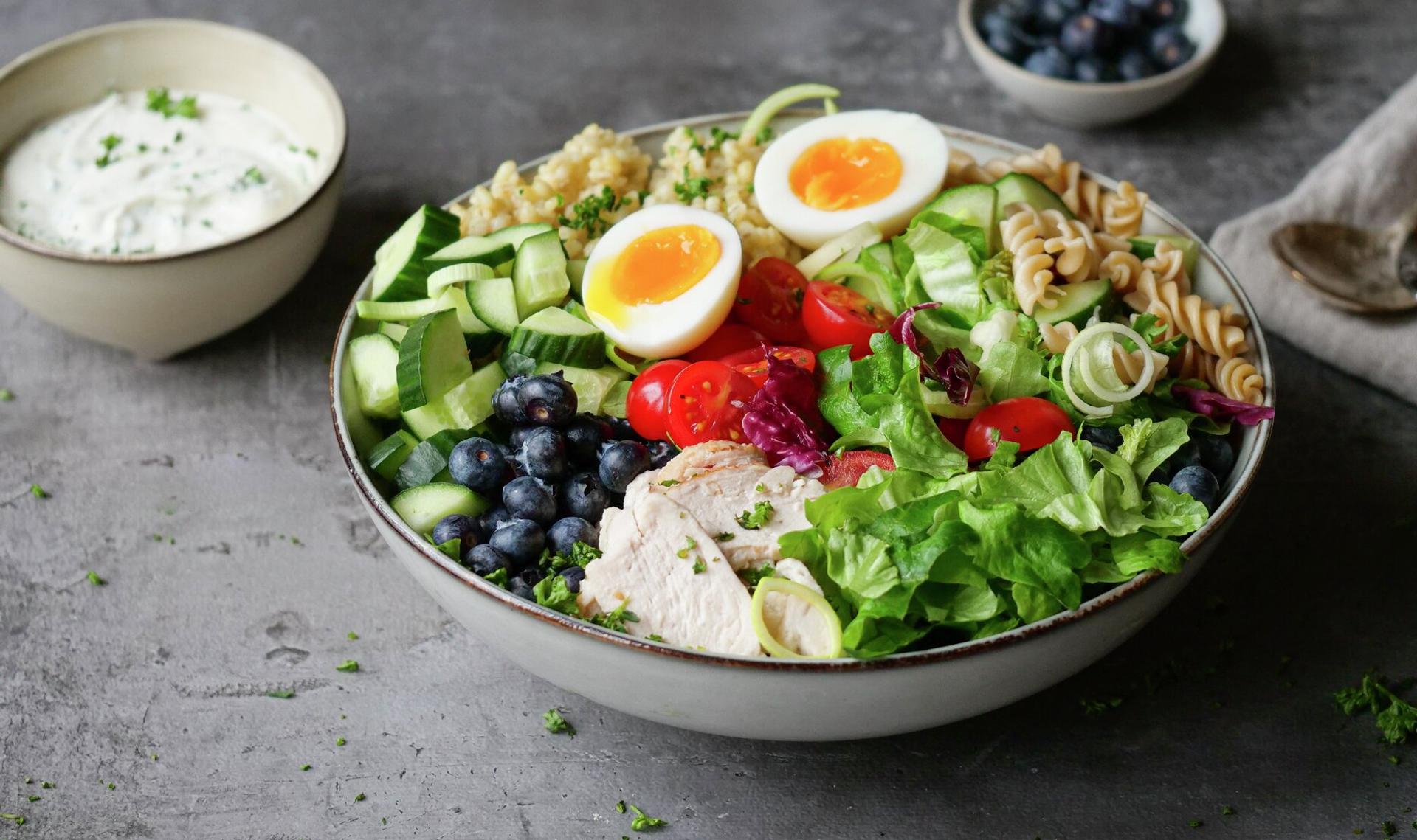 A vibrant healthy meal bowl with sliced chicken, soft-boiled eggs, blueberries, mixed greens, pasta, and grains, served with a side of white dressing.