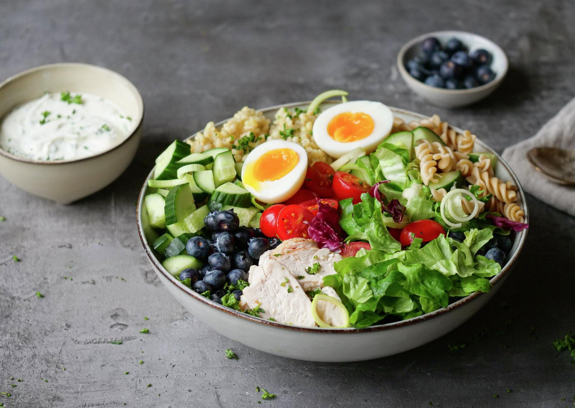 A colorful Buddha bowl with chicken, soft-boiled eggs, mixed greens, cucumber, tomatoes, blueberries, pasta, and a creamy dressing on the side.