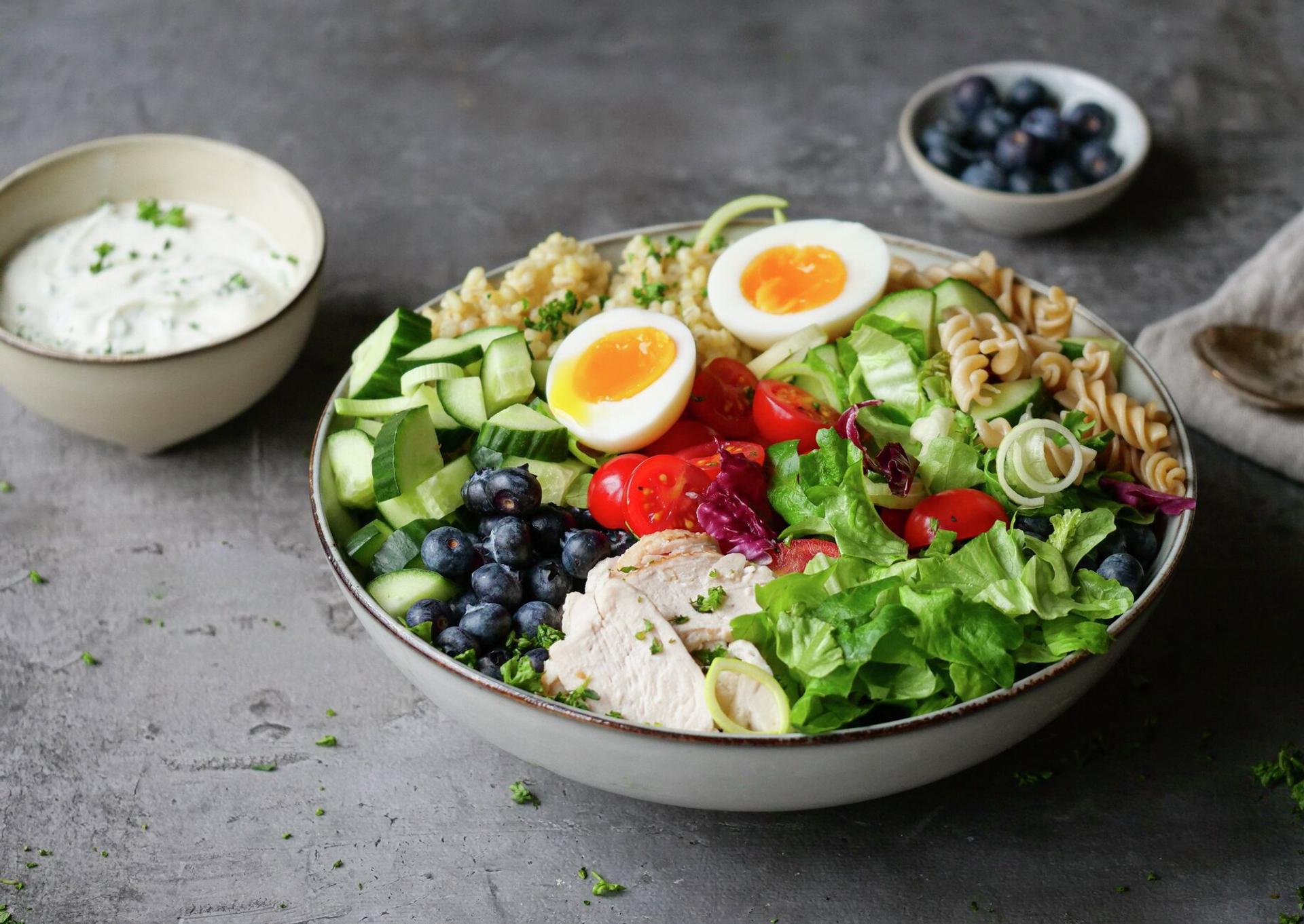 A colorful buddha bowl with chicken, soft-boiled eggs, blueberries, cucumber, tomatoes, lettuce, pasta, and grains, with a side of dressing.