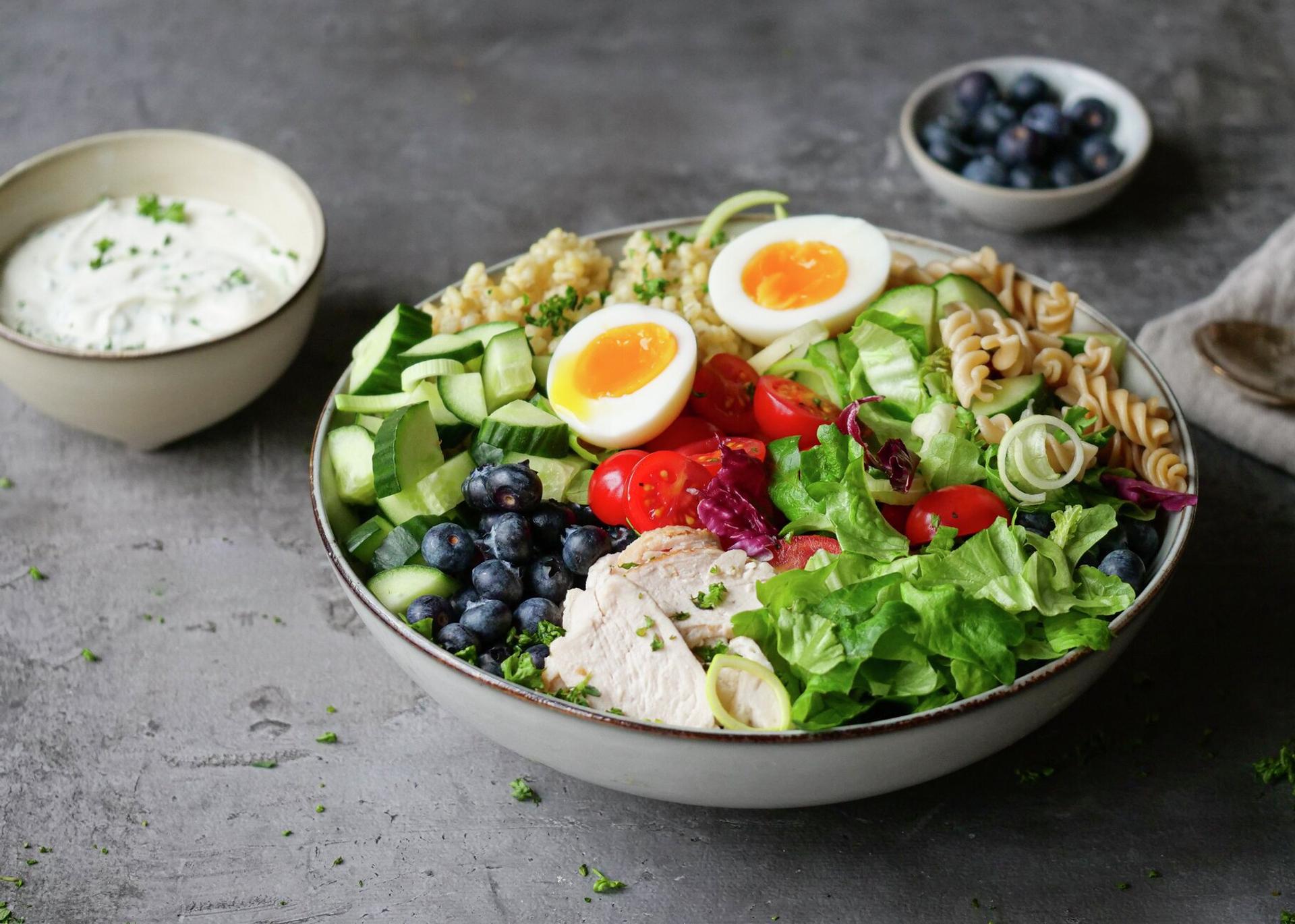 A colorful salad bowl filled with chicken, two soft-boiled eggs, blueberries, pasta, mixed greens, cucumbers, and tomatoes, with a side of white dressing.