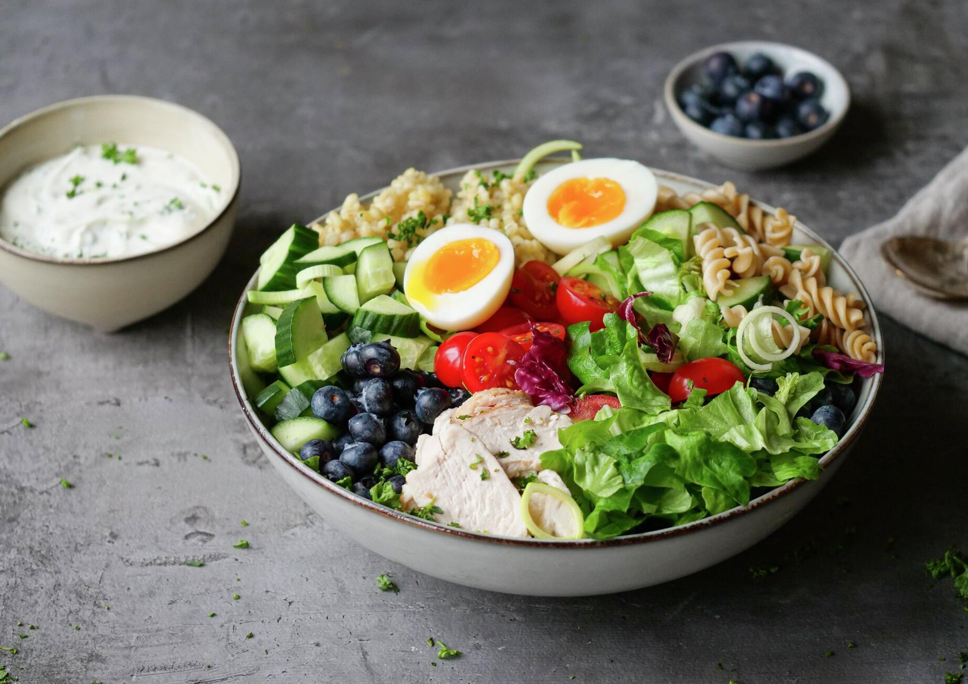 A colorful salad bowl with chicken, soft-boiled eggs, blueberries, cucumber, tomatoes, greens, pasta, and quinoa, with a side of dressing.
