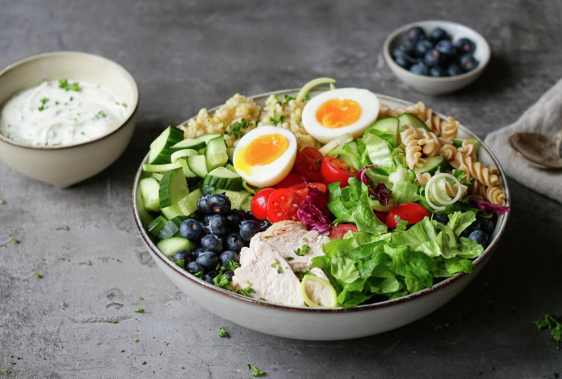A colorful grain bowl with sliced chicken, soft-boiled eggs, blueberries, cucumber, tomatoes, mixed greens, pasta, and quinoa, served with a side of white dressing.