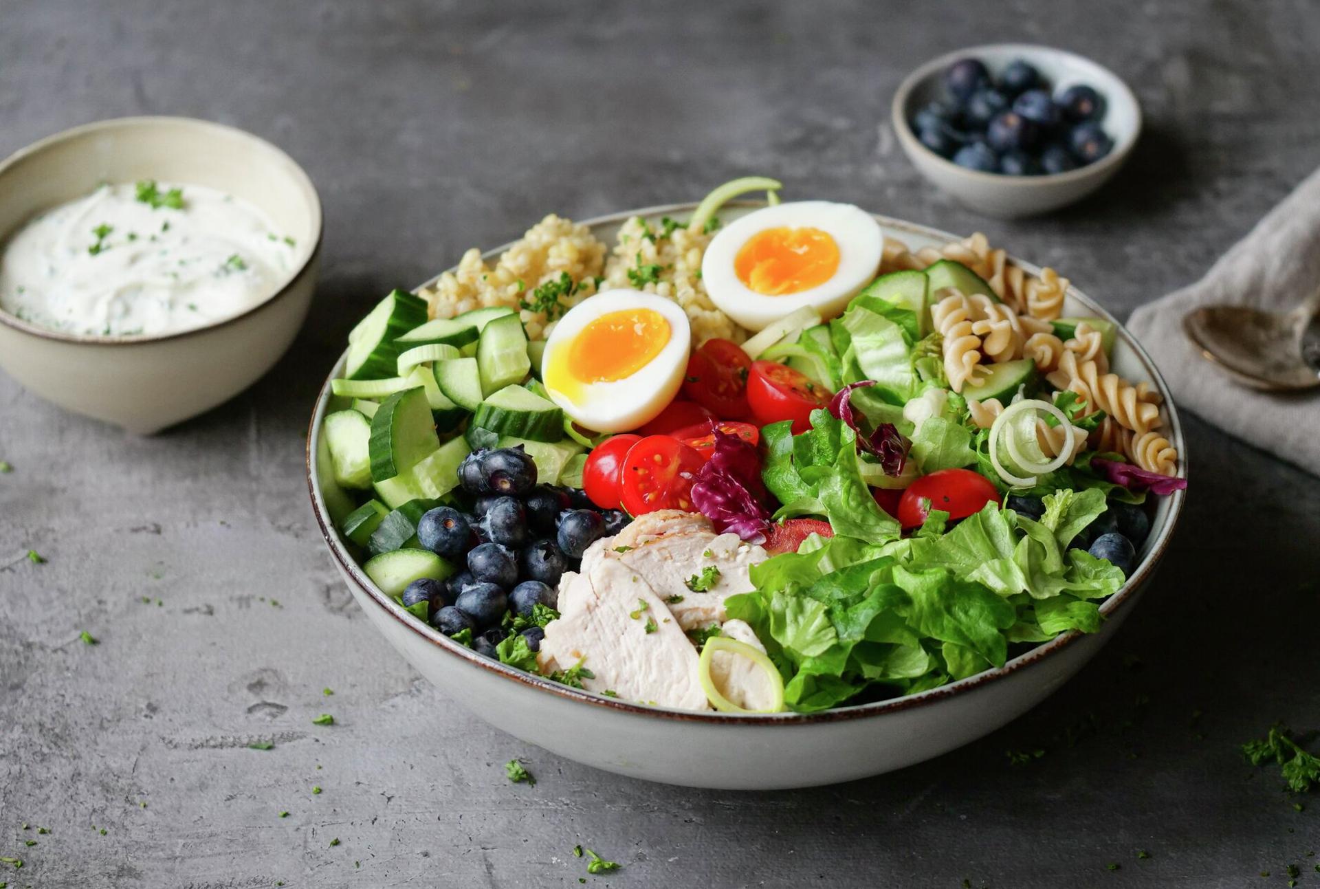 A colorful meal bowl with sliced chicken, two halved soft-boiled eggs, blueberries, cucumber, tomatoes, lettuce, pasta, and a grain, served with a side of white sauce.