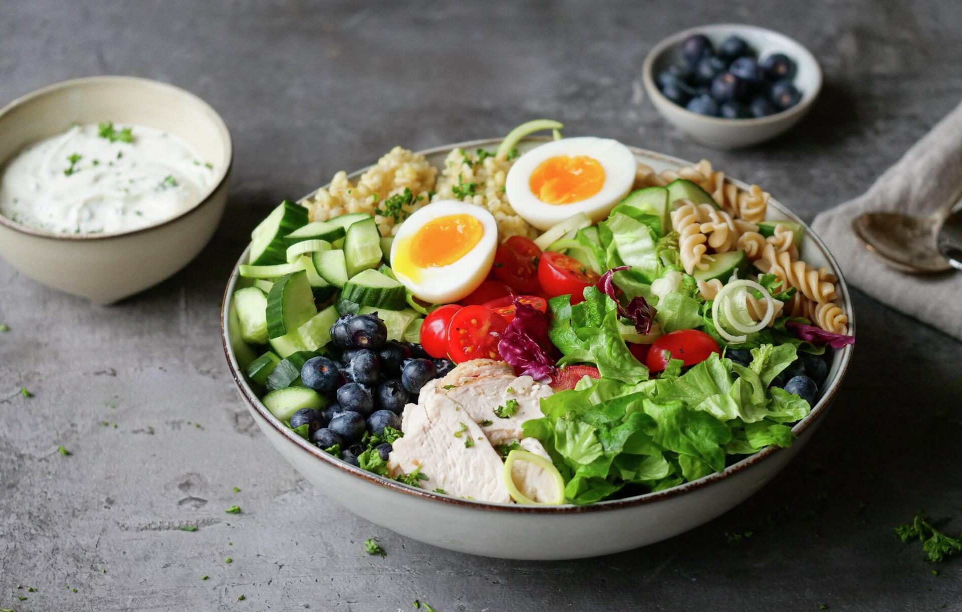 A vibrant Buddha bowl filled with chicken, halved soft-boiled eggs, blueberries, cucumber, tomatoes, mixed greens, pasta, and grain, served on a grey surface with a side of dressing.