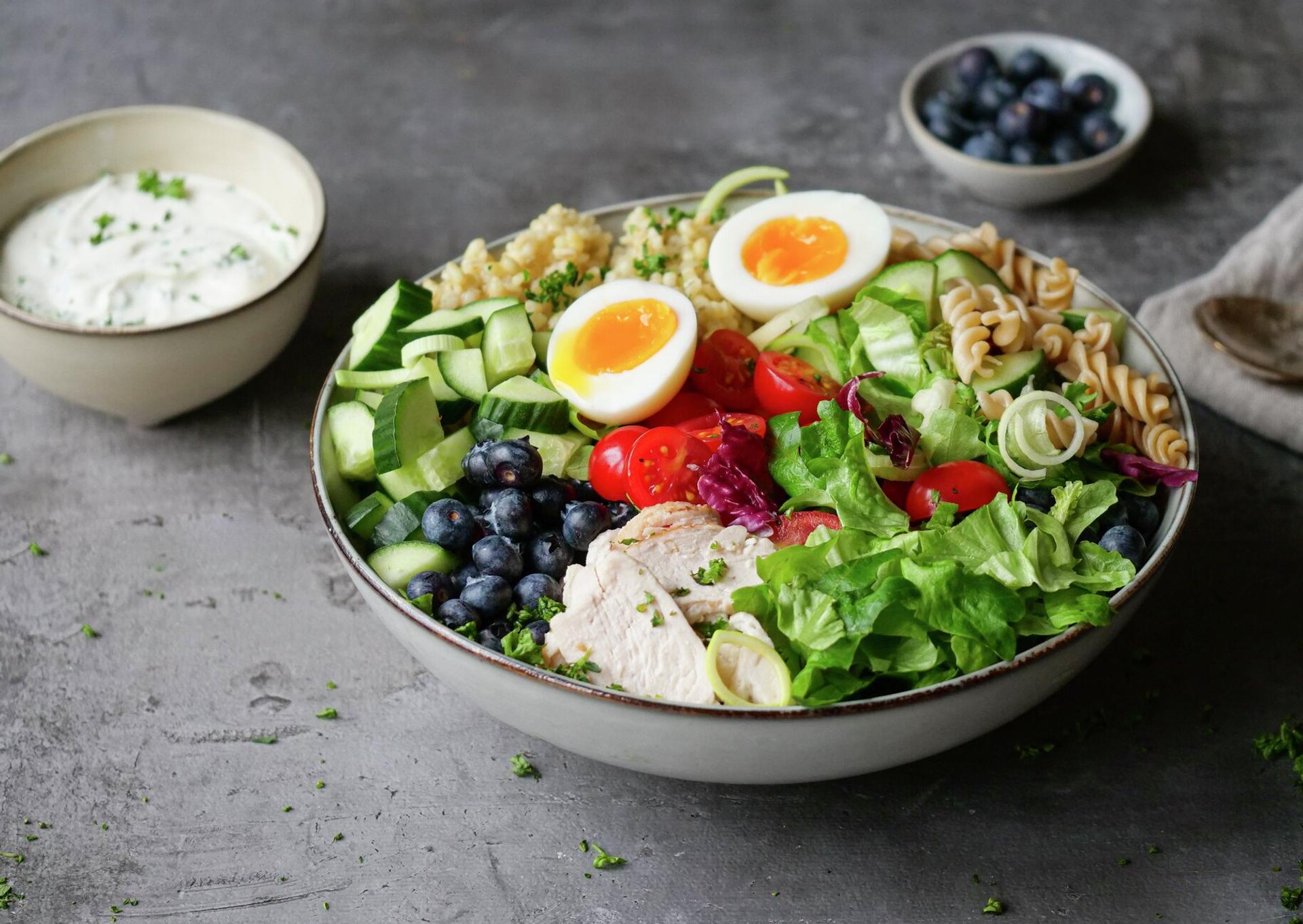 A vibrant salad bowl with sliced chicken, two soft-boiled eggs, blueberries, fresh vegetables, grain, and pasta, served with a side of white dressing.