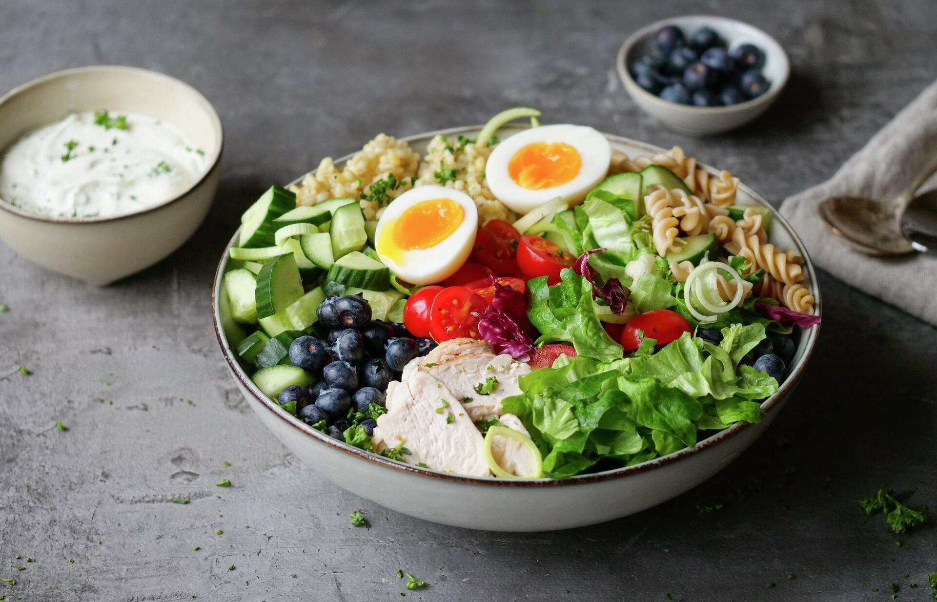 A colorful protein bowl with sliced chicken, soft-boiled eggs, blueberries, mixed greens, pasta, grains, and a side of white sauce.