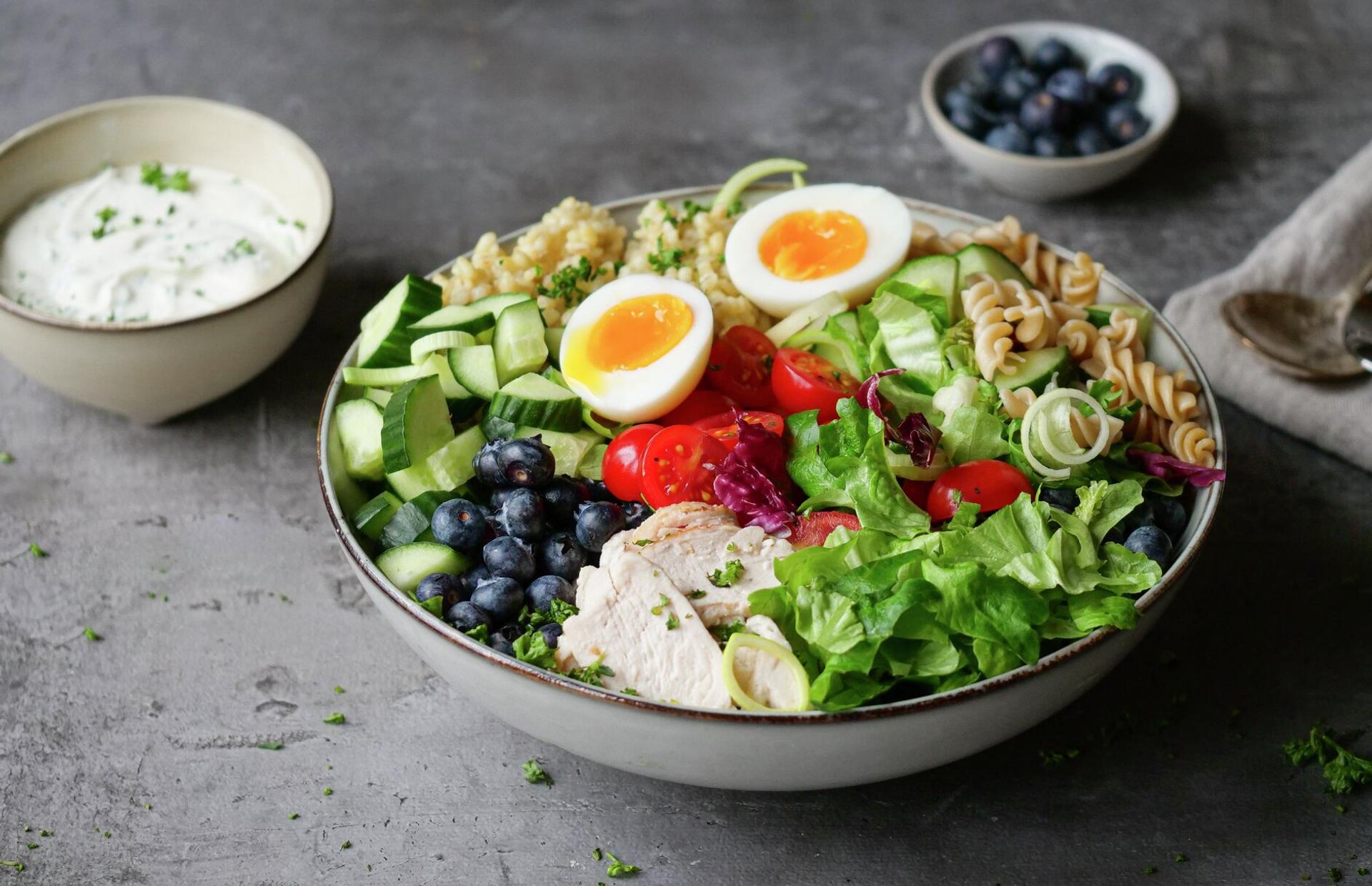 A vibrant meal bowl with sliced chicken, two soft-boiled eggs, blueberries, cucumber, tomatoes, mixed greens, quinoa, and pasta, with a side of creamy herb dressing.