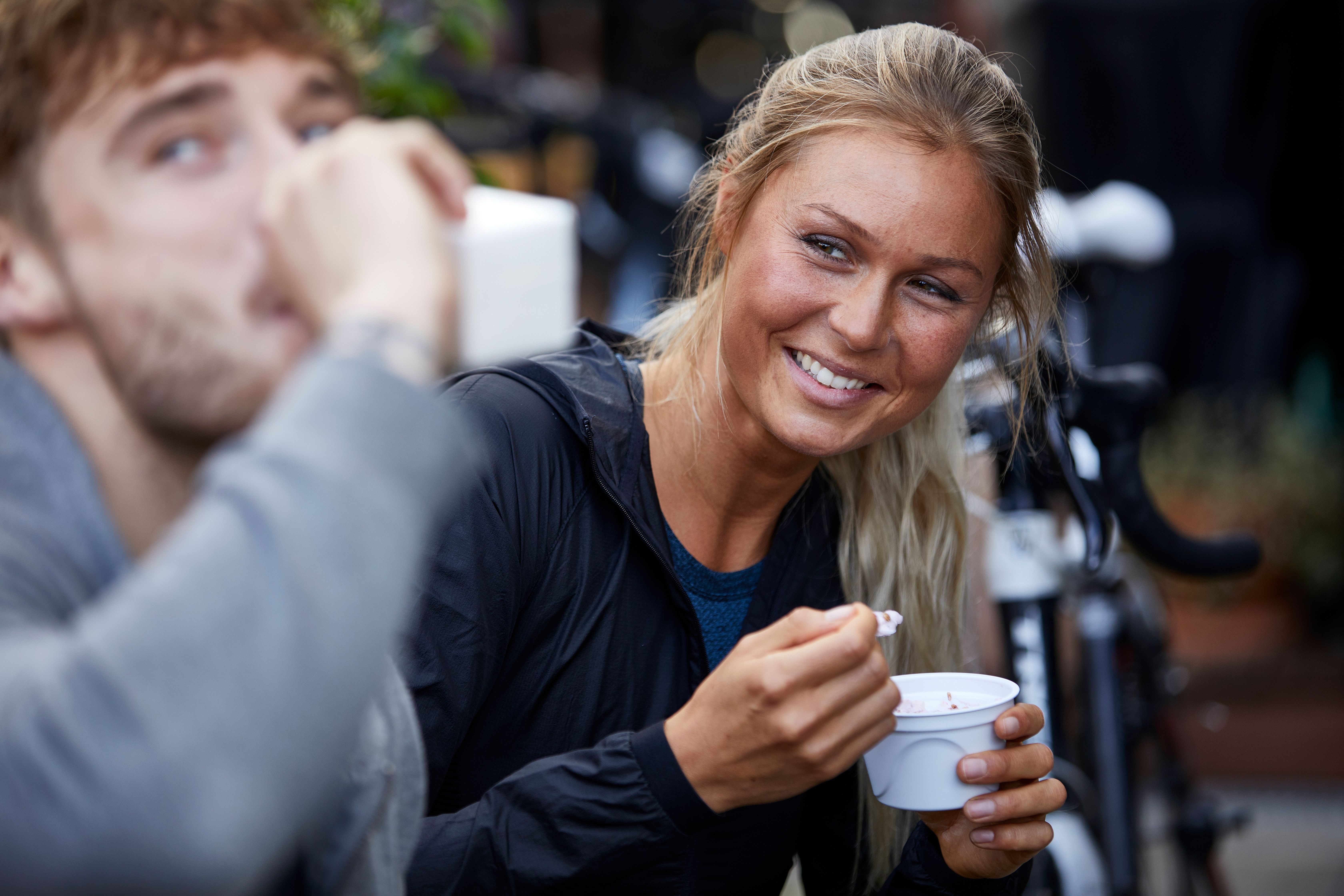 A smiling woman eats from a cup, with a blurred man drinking in the foreground.