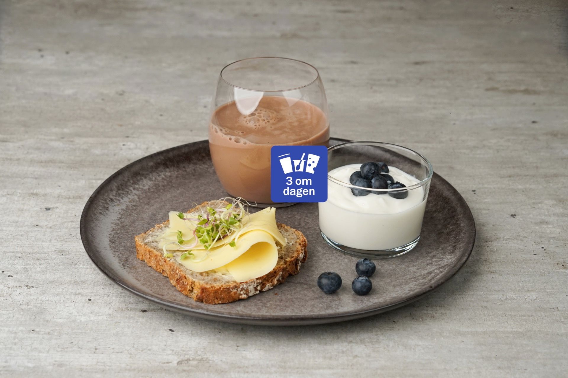 Grå tallerken med brødskive med gulost og spirer, yoghurt ed blåbær og et glass sjokolademelk