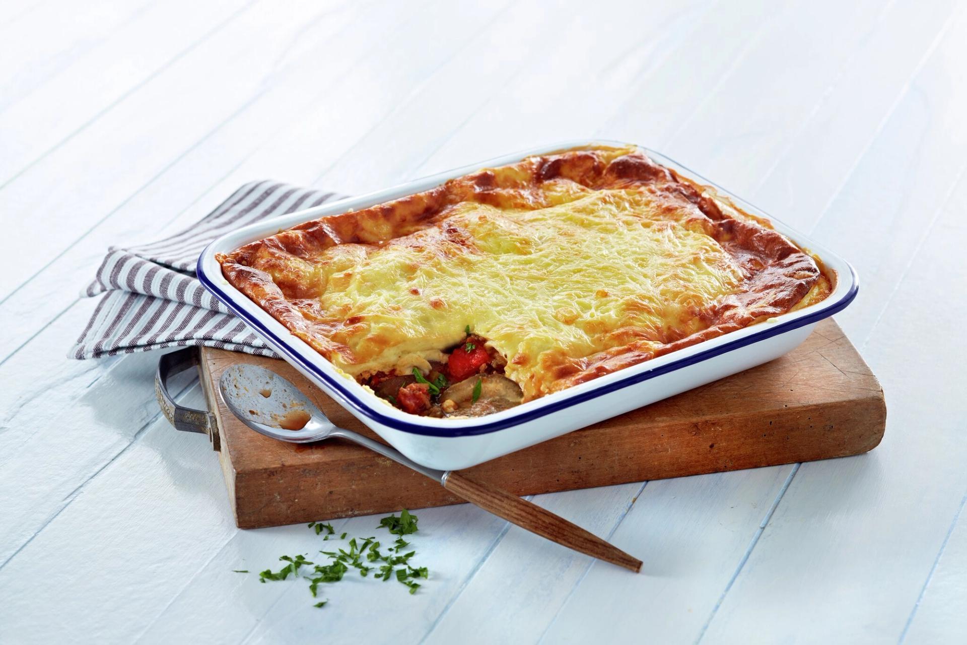 A golden-brown baked casserole in a white dish, a portion scooped out to reveal colorful vegetables, with a spoon resting inside on a wooden board.