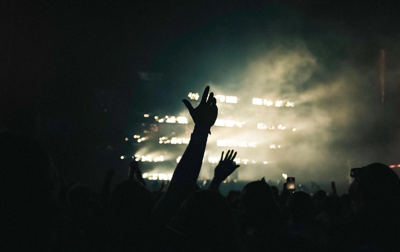 Silhouetted concert crowd with raised hands in front of a stage glowing with white lights and haze.