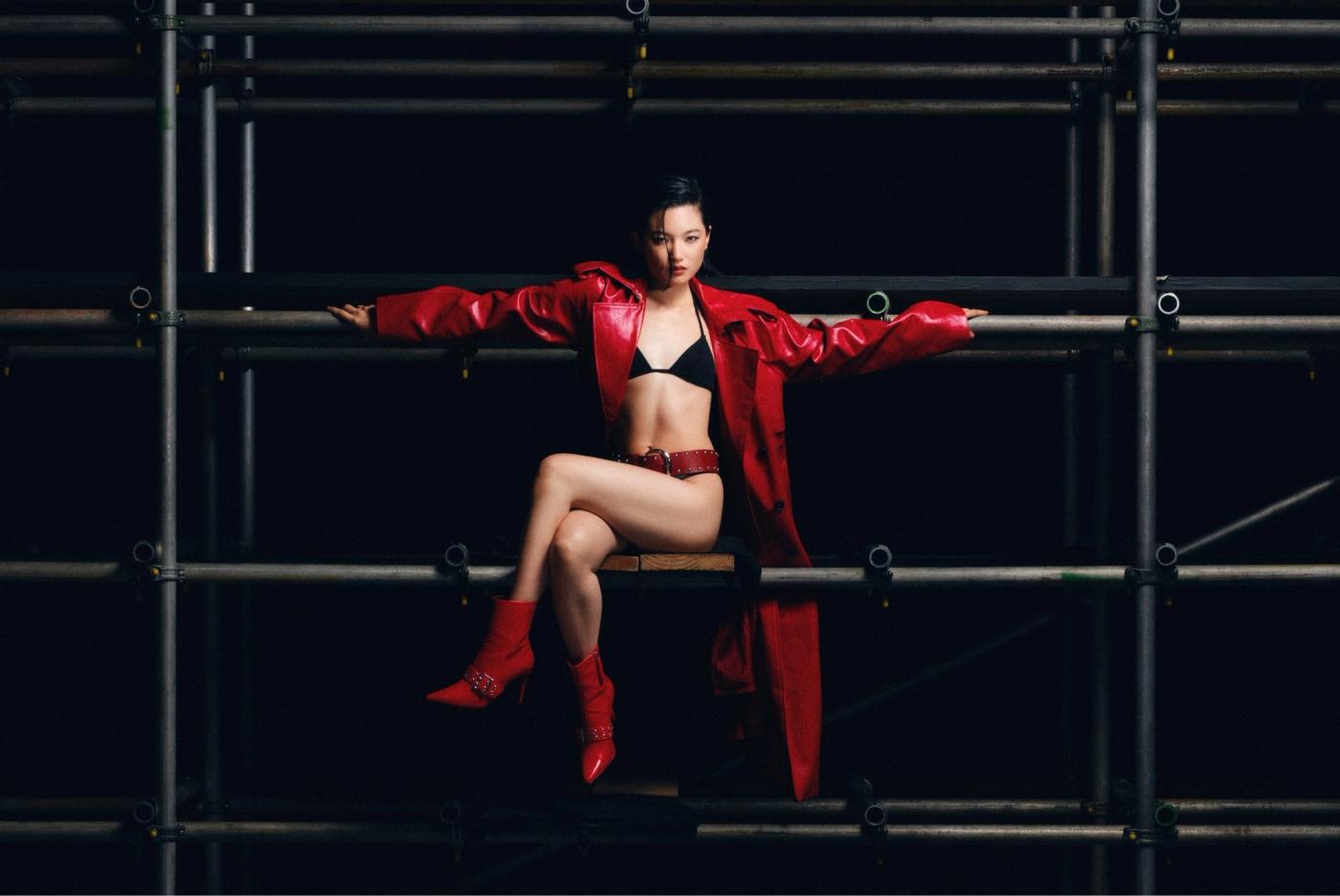 Chloe Qisha in a red leather coat and red boots, seated on scaffolding against a black background