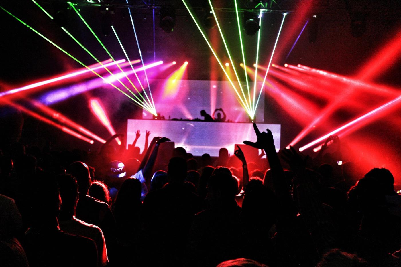 A club crowd seen from behind with hands raised under green, pink and red laser beams, a lit DJ booth in the distance.