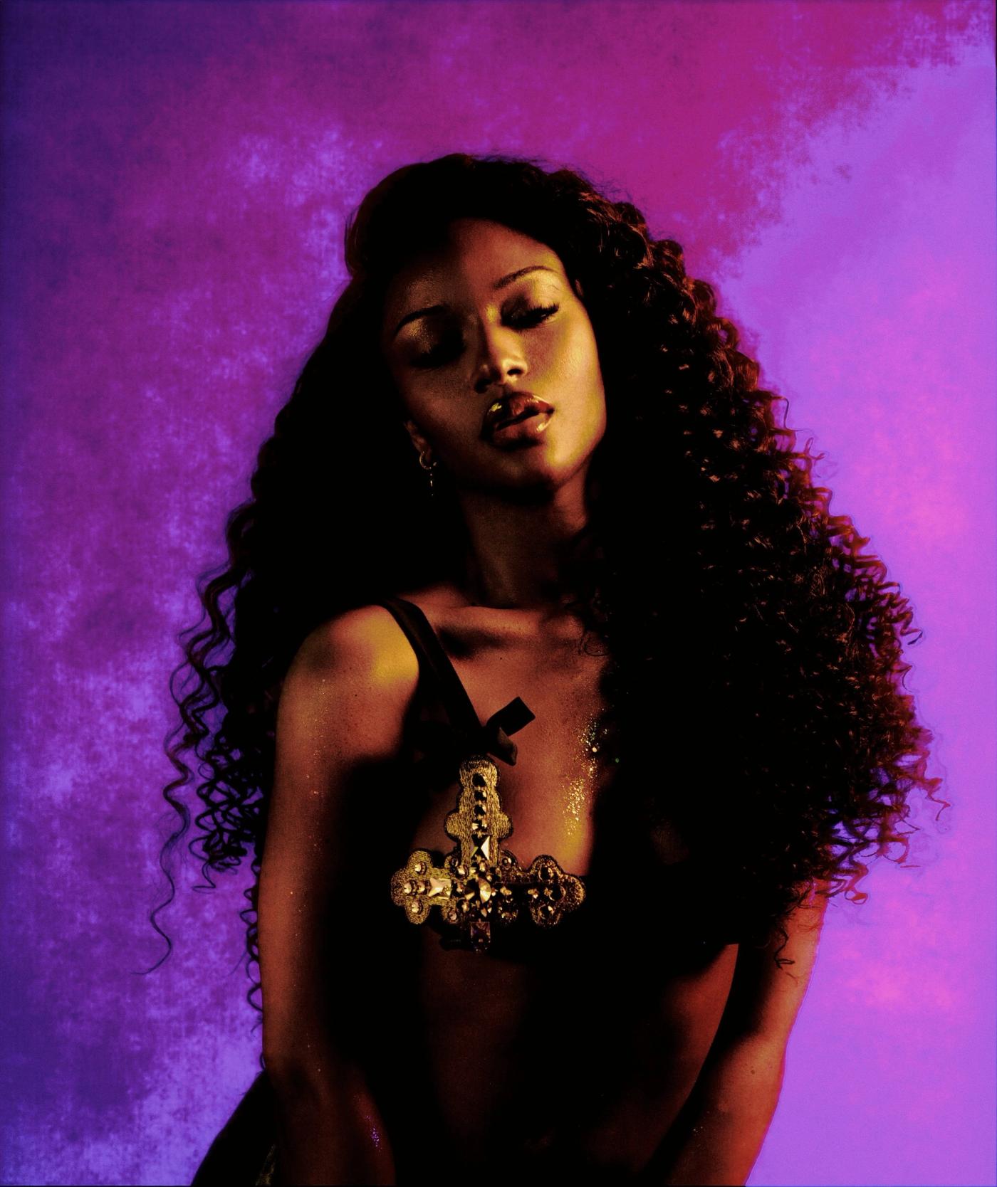 Ayra Starr with long curly hair and closed eyes, wearing a jeweled gold cross, against a purple and pink backdrop