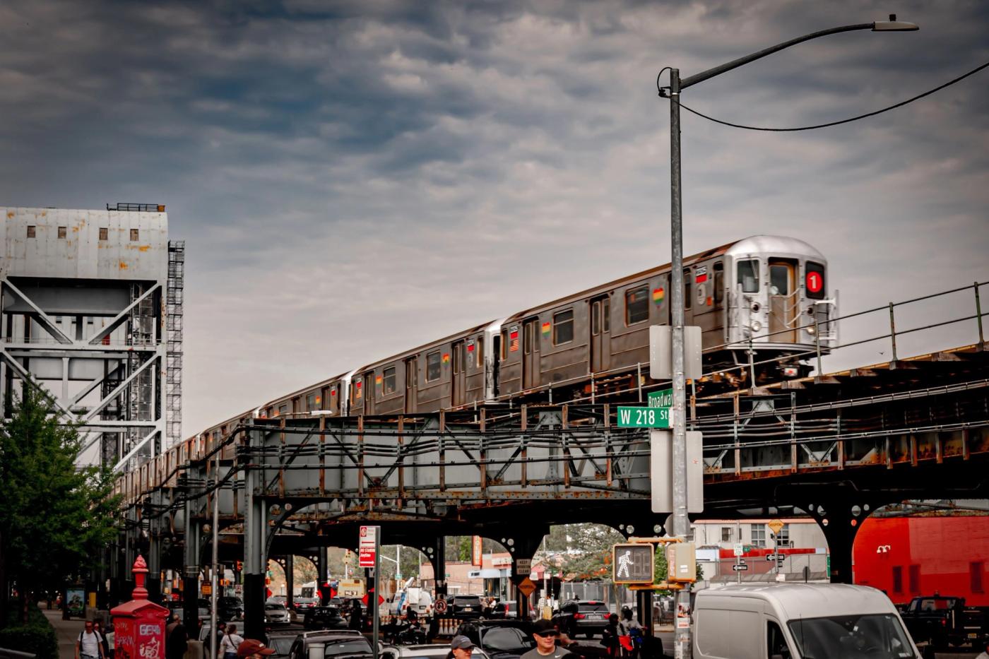A 1 train crosses the elevated tracks above Broadway at West 218th Street in Manhattan under a cloudy sky, with traffic below.