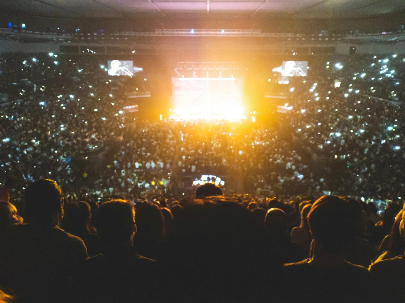 Crowd in a packed indoor arena holding up phone lights toward a brightly lit stage