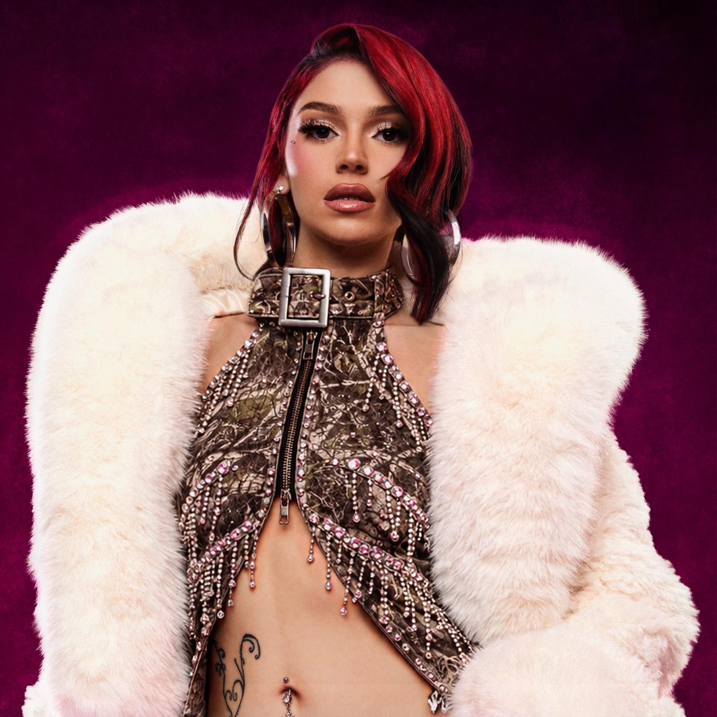 cieratherapper with red and black bob-length hair and large silver hoop earrings, in a camo halter top with rhinestone fringe and a cream faux-fur coat, against a magenta background