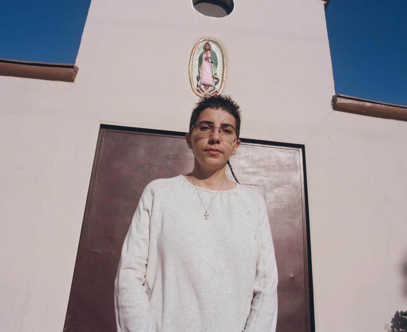 St. Panther in glasses, a gray sweatshirt and a cross necklace stands before a white building with a brown door and a Virgin of Guadalupe image above