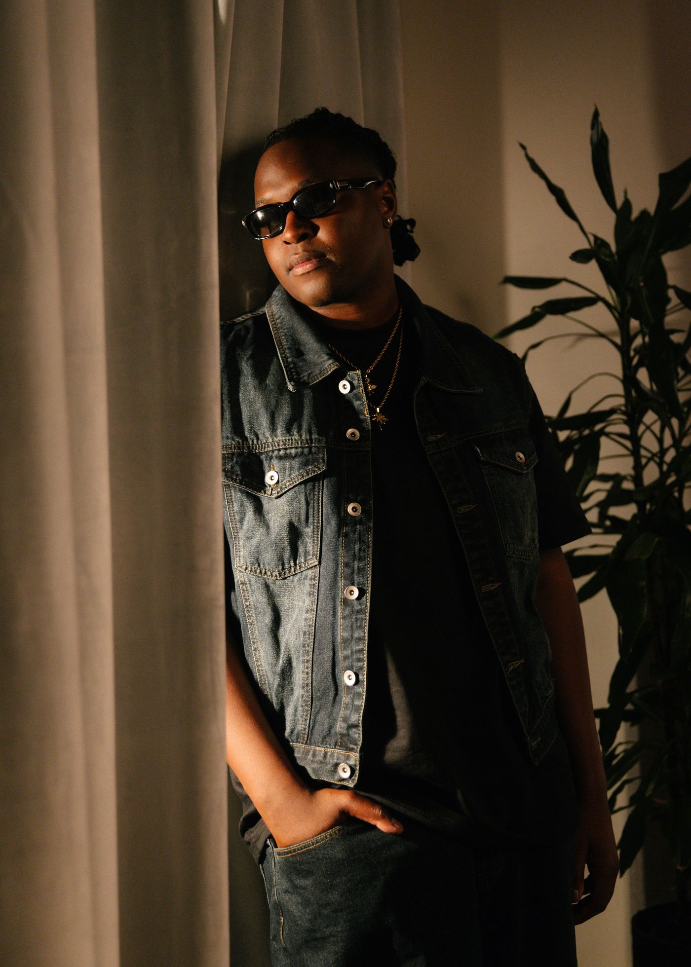 Kuzi Cee in sunglasses and a denim vest, leaning beside a curtain in warm light