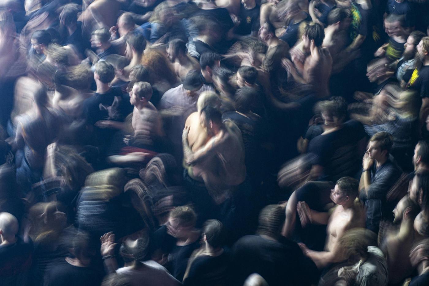 Overhead long-exposure shot of a packed, motion-blurred concert crowd in a dark venue, several people shirtless.