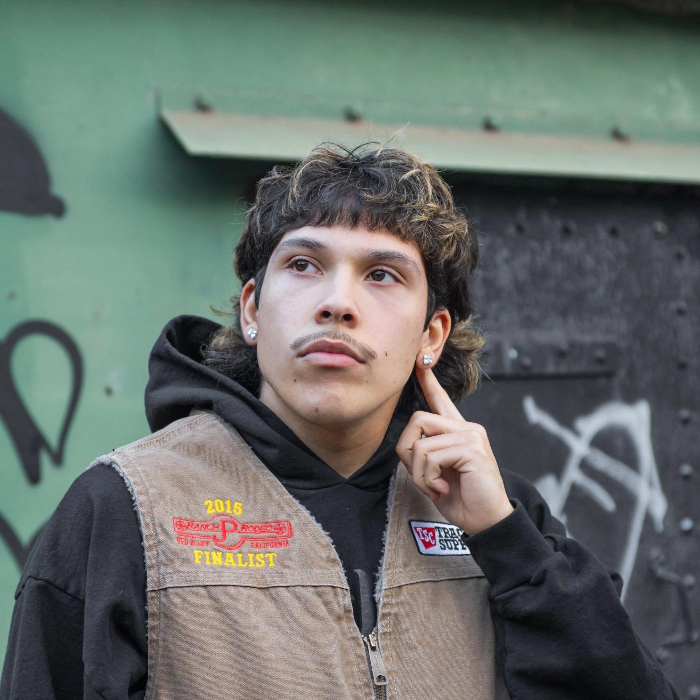 SOOS in a tan work vest with a 2015 Finalist patch over a black hoodie, finger to his ear, in front of a green and black wall with graffiti