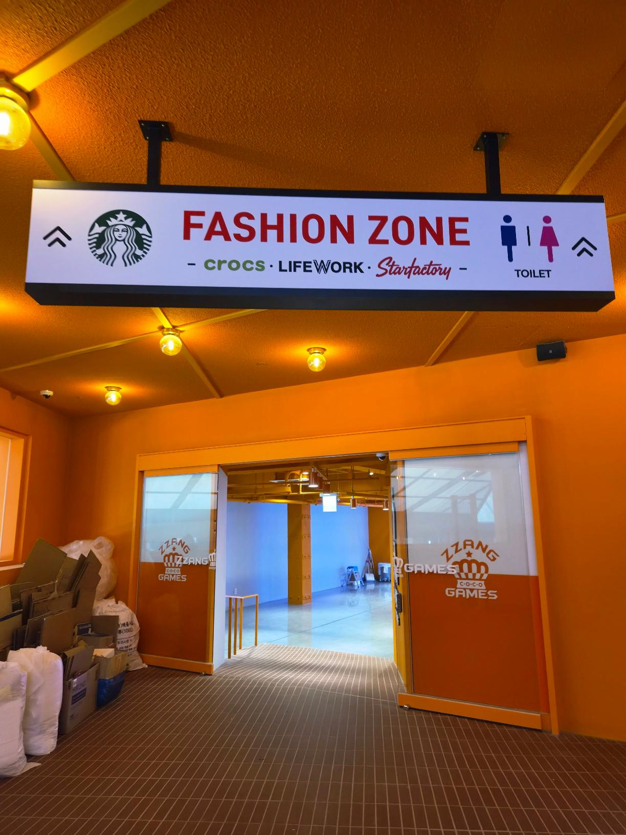 An orange-walled corridor with a modern illuminated sign reading Fashion Zone pointing toward Starbucks, Crocs, and Lifework.