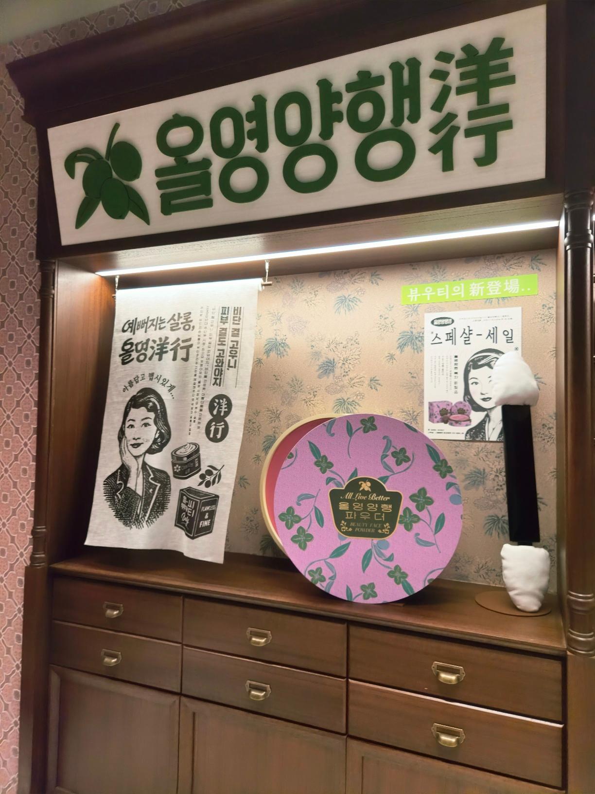 A vintage beauty vanity display showcasing an oversized pink floral powder tin and retro Korean cosmetic posters.