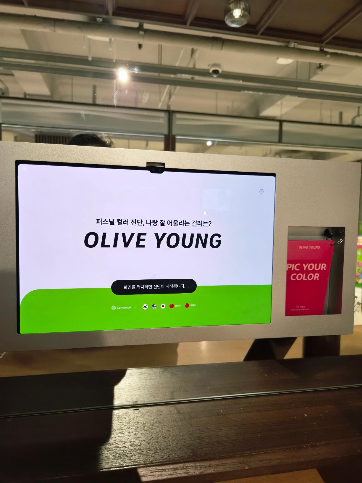 A sleek silver personal color diagnosis kiosk displaying a white start screen that reads Olive Young in bold.