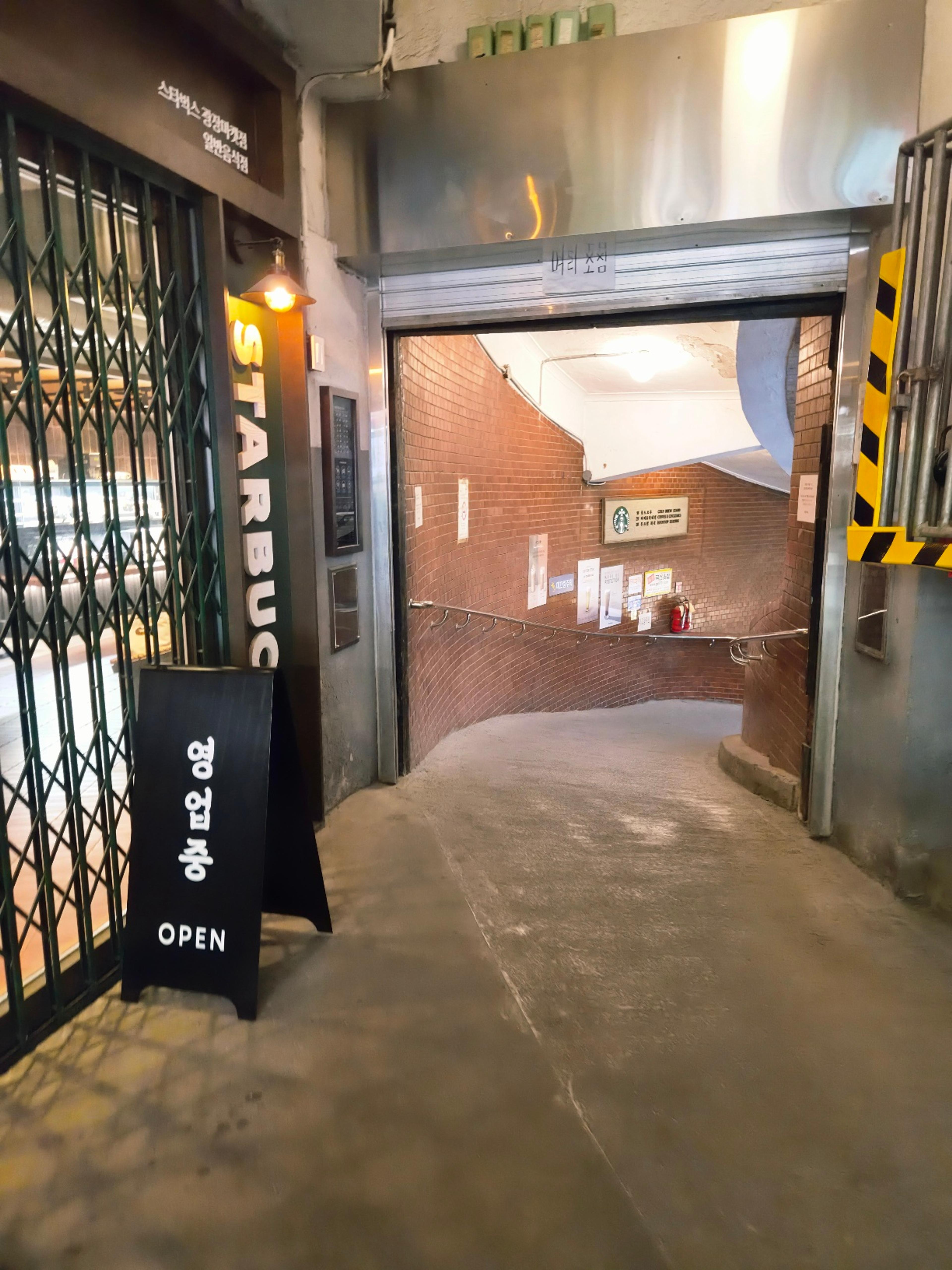 A minimalist entrance to a Starbucks coffee shop located down a rustic, curved red brick tunnel hallway.