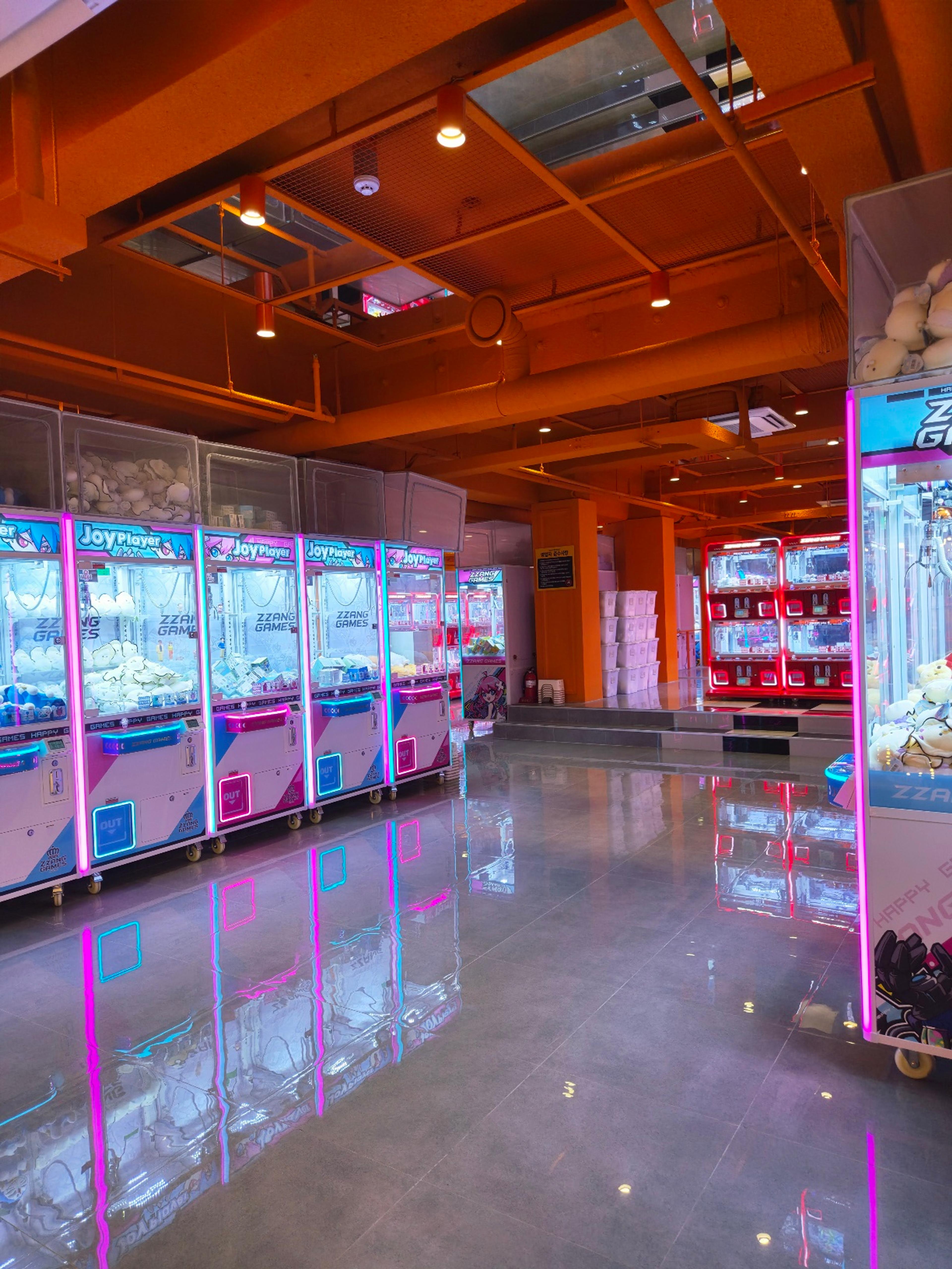 A wide interior shot of a spacious, modern arcade lined with glowing neon pink and blue plushie claw machines.