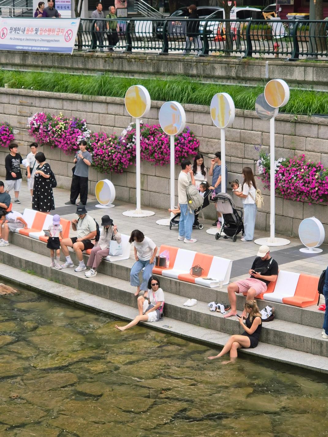 People sitting on orange-and-white benches along the Cheonggyecheon stream in downtown Seoul, with some dipping their feet into the shallow water and colorful flower beds lining the stone wall behind them