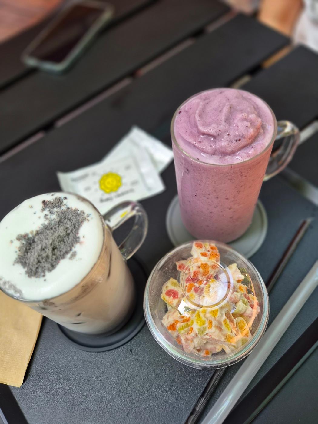 Close up of a Black Sesame Latte, a Blueberry Smoothie, and a colorful cereal marshmallow cookie on a dark table