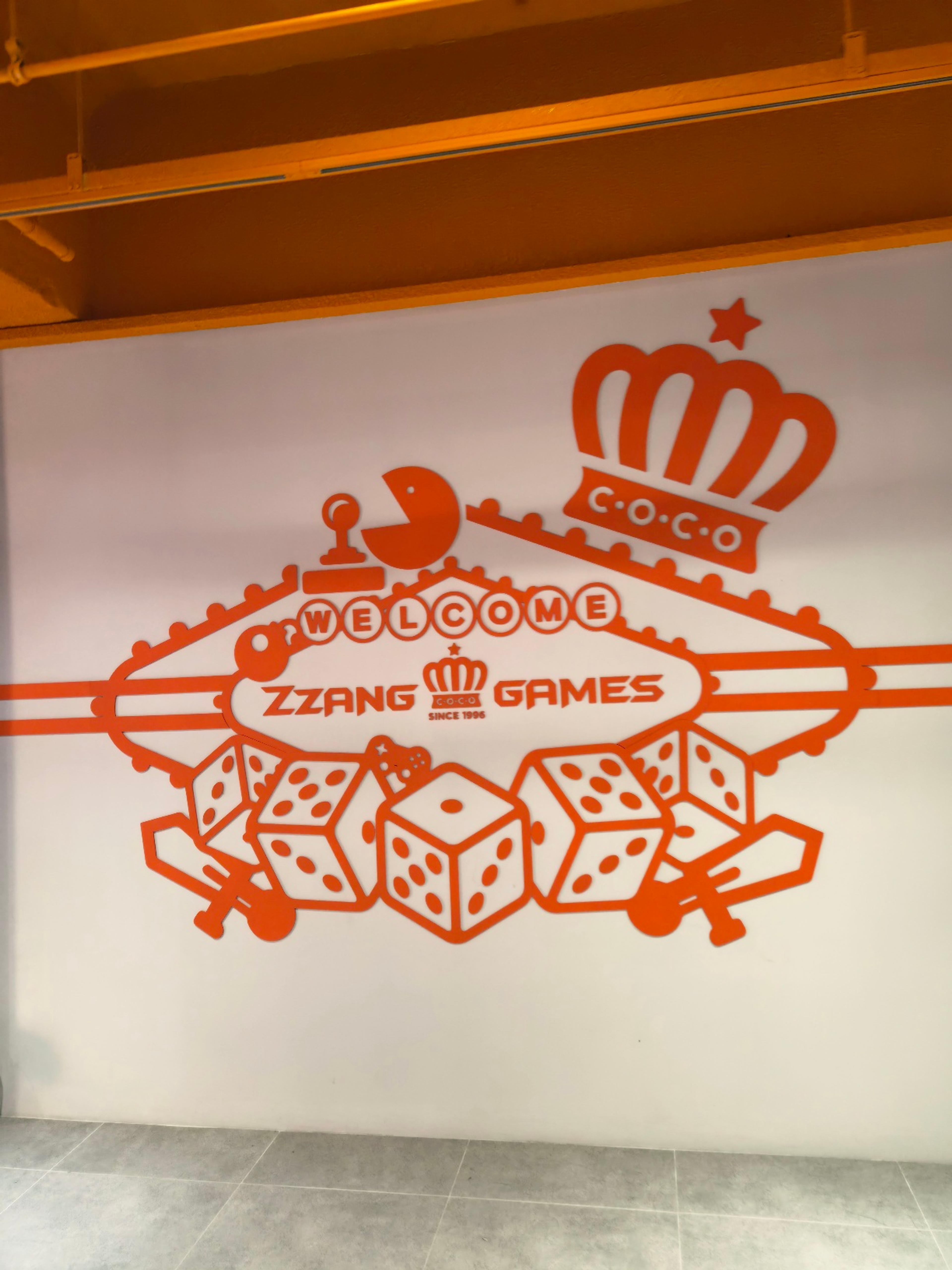 A vibrant orange and white wall graphic welcoming visitors to Zzang Games with illustrations of dice, Pacman, and a crown.