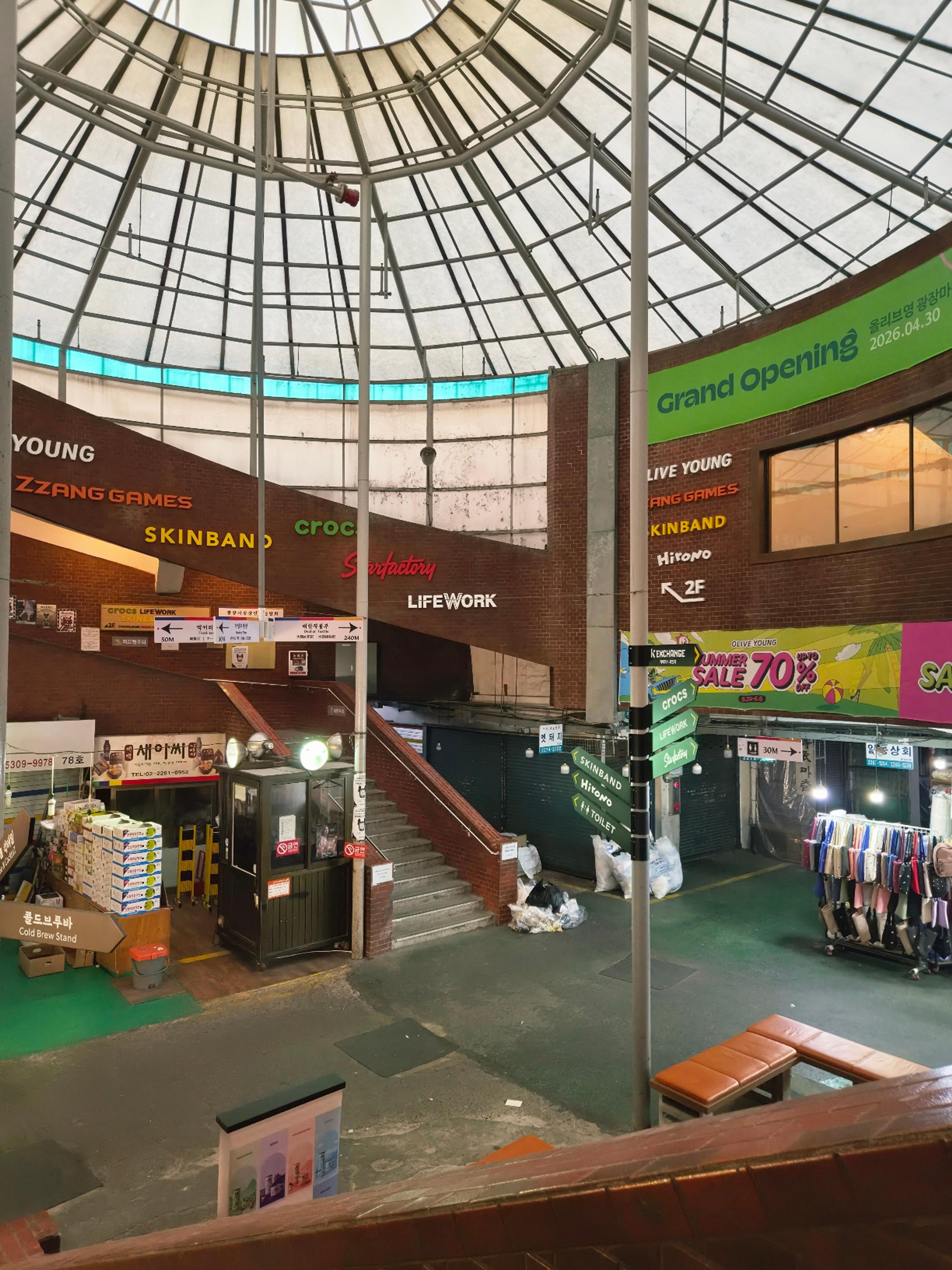 A wide shot of the Gwangjang Market central dome featuring retail directory signs for Olive Young, Zzang Games, and a green Grand Opening banner.
