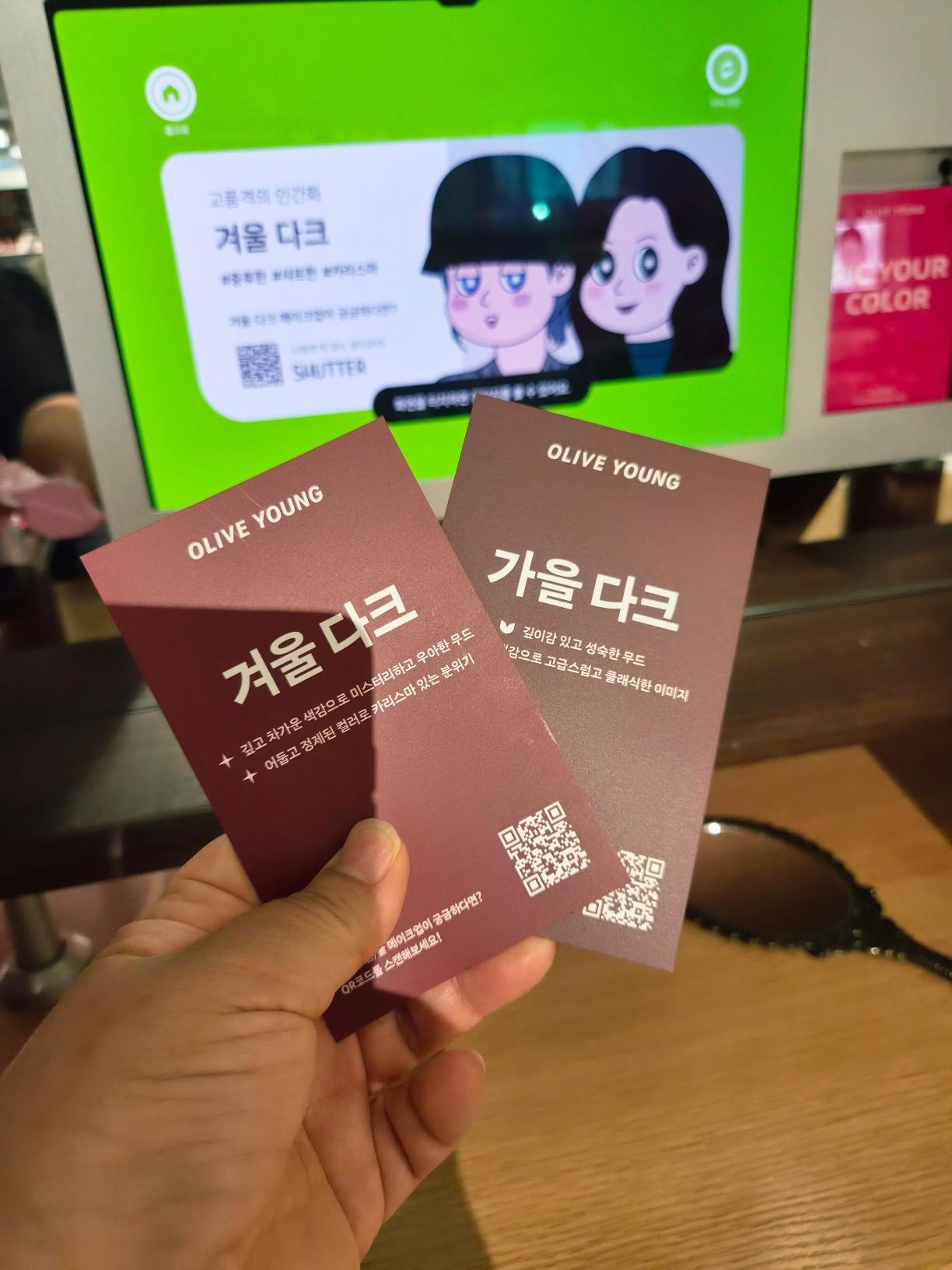 A hand holding two premium personal color result cards from Olive Young reading Winter Dark and Autumn Dark.