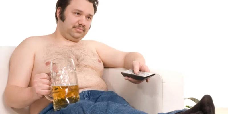 Overvægtig mand, der sidder passivt foran fjernsynet med et stort krus øl.