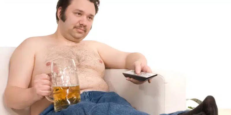 Överviktig man som sitter passivt framför TV:n med en stor mugg öl.