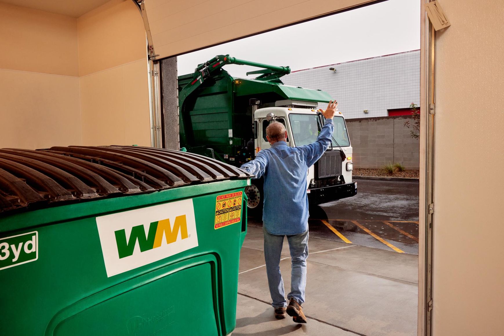 A Waste Management employee directs a garbage truck backing into a bay with a green dumpster.