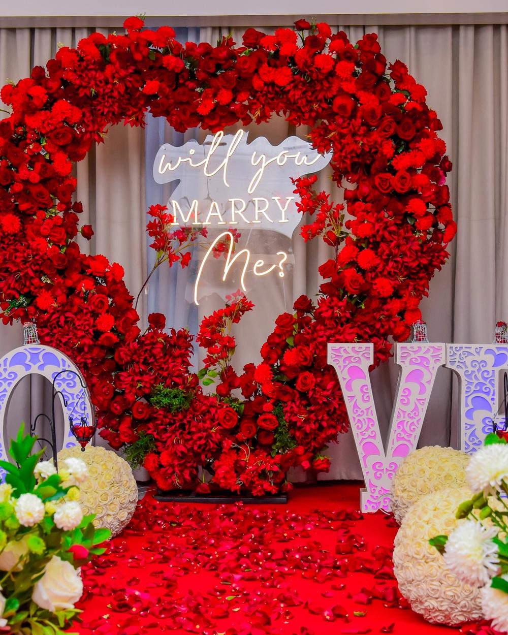 Rose-covered heart-shaped arch with a neon 'will you marry me?' sign, proposal setup