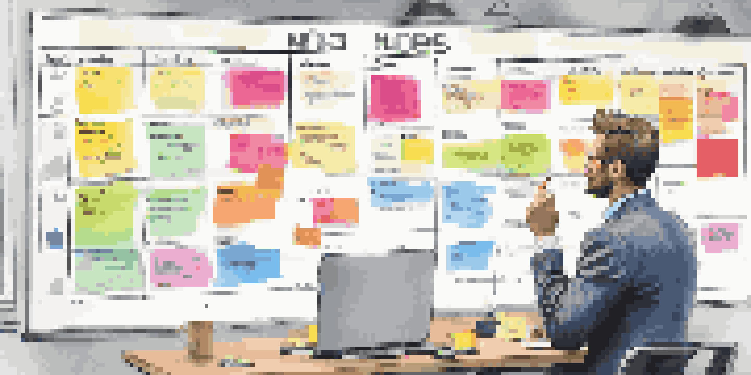 A person in a modern office pointing at a colorful marketing timeline on a wall calendar, with sticky notes and a laptop nearby.