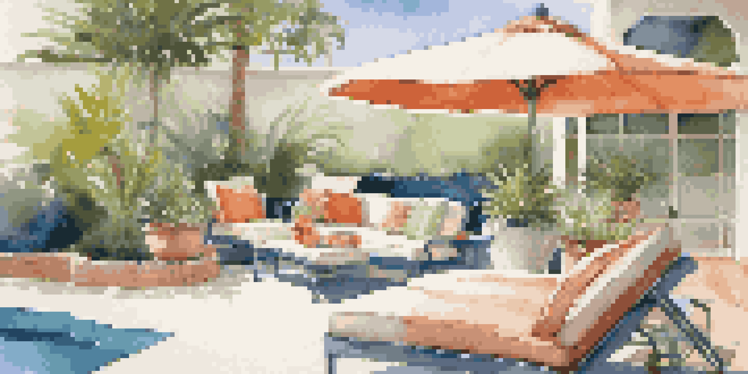 A picturesque poolside lounge area with colorful cushions, a small table, and a shade umbrella surrounded by greenery and a clear blue pool.