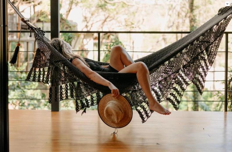 Woman resting in a black macramé hammock on a wooden treehouse deck, hat in hand, jungle behind