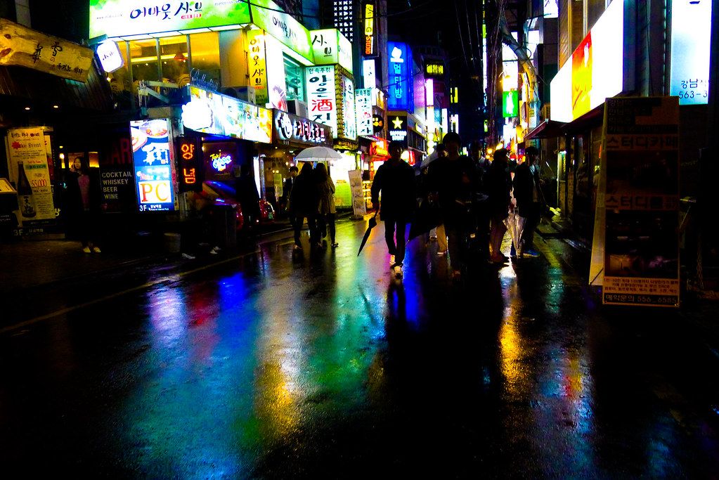 A rainy night in Seoul, with neon reflections on wet streets and people carrying umbrellas.