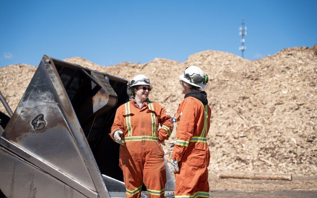 Two workers in orange safety overalls and helmets engage in conversation at a wood chip processing site. A large pile of wood chips is visible in the background under a clear blue sky.