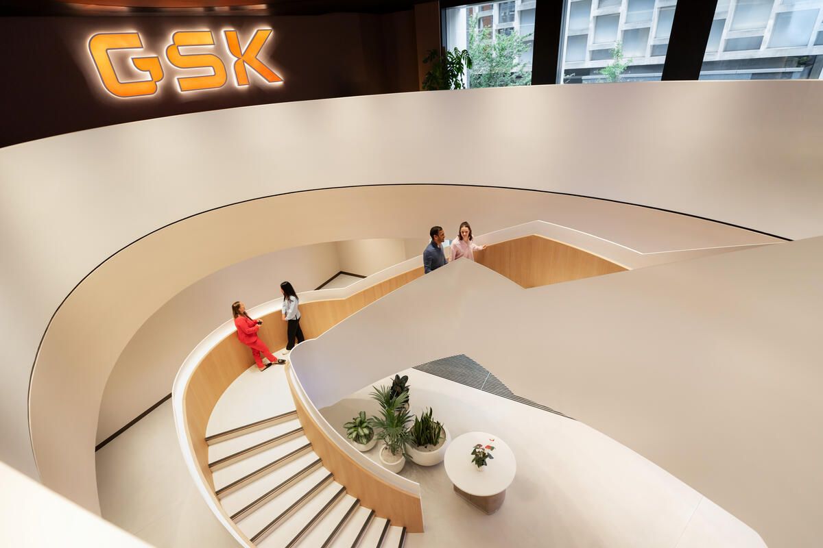 The image depicts an interior view of a modern corporate building featuring a spiral staircase. Three individuals are seen interacting, while a large 'GSK' logo is prominently displayed on the wall.
