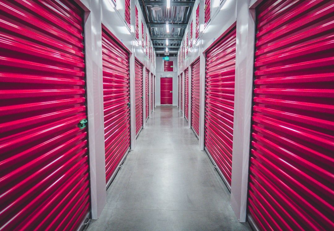 This image shows a neatly arranged hallway of a storage facility, featuring bright red storage units on either side. The corridor is well-lit, clean, and has a polished concrete floor.