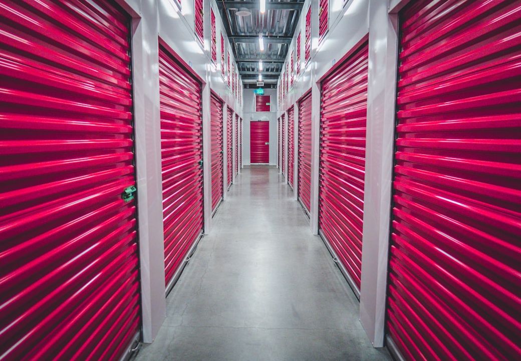 This image shows a neatly arranged hallway of a storage facility, featuring bright red storage units on either side. The corridor is well-lit, clean, and has a polished concrete floor. — Credit: Photo by JOSHUA COLEMAN on Unsplash