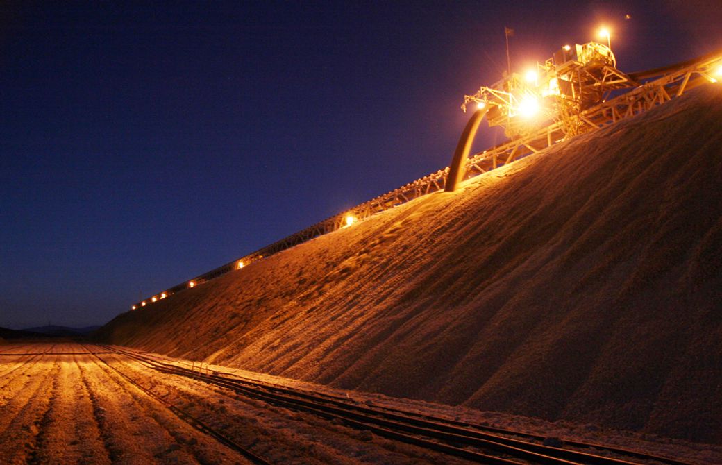 The image depicts an industrial scene at night, featuring a large conveyor system illuminated by powerful lights. The conveyor is used for transporting materials from a high mound of stacked material, likely aggregate or grain, to another location.