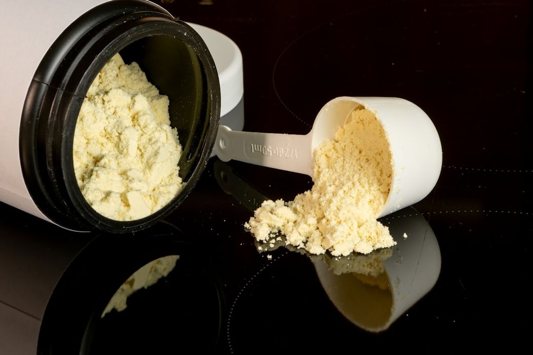 The image depicts a partially opened container of powder supplement with a measuring scoop beside it. Some of the powder is spilled on a reflective surface, creating a visually striking composition. — Credit: Photo by Alex Saks on Unsplash
