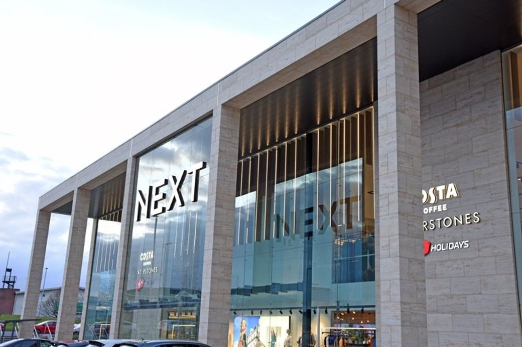 The image shows the exterior of a NEXT retail store, highlighting large glass windows and a modern architectural design. Adjacent to the store, there is a Costa Coffee shop, indicating a shopping environment.